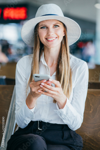 Young woman wearing white shirt and hat holding smartphone while sitting in airport waiting area