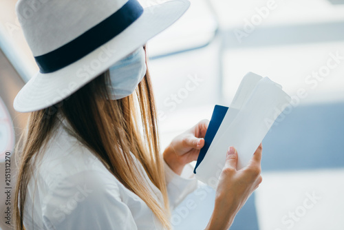 Woman wearing wide-brimmed hat and face mask checks passport and boarding pass while waiting for flight at airport terminal