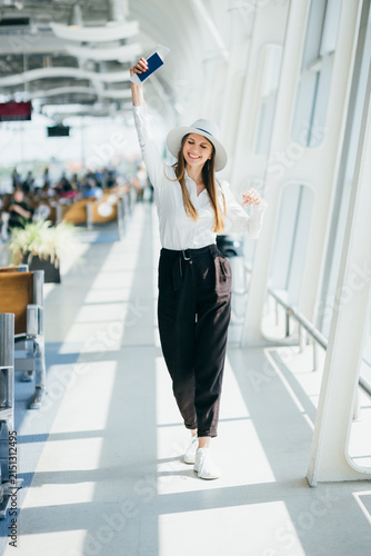 Bild auf Leinwand Young woman in stylish outfit holding passport and boarding pass aloft in airpor