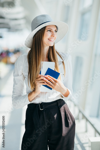 Woman wearing hat and white shirt holding passport and ticket in airport terminal