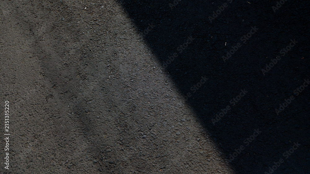 Fototapeta premium Dark rough asphalt road surface texture split by diagonal sunlight shadow.