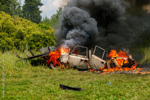 Old car burning in a green grassy field with bright orange flames and thick black smoke rising into the sky against a background of trees and bushes on a summer day