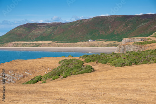 A summer view over Rhosilli Bay, with sun-baked grassland in the foreground overlooking the sandy beach and blue sea beneath a bright, cloudless sky.