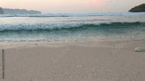 Gentle waves rolling onto a sandy beach at sunset with a distant island landscape and a seashell in the foreground