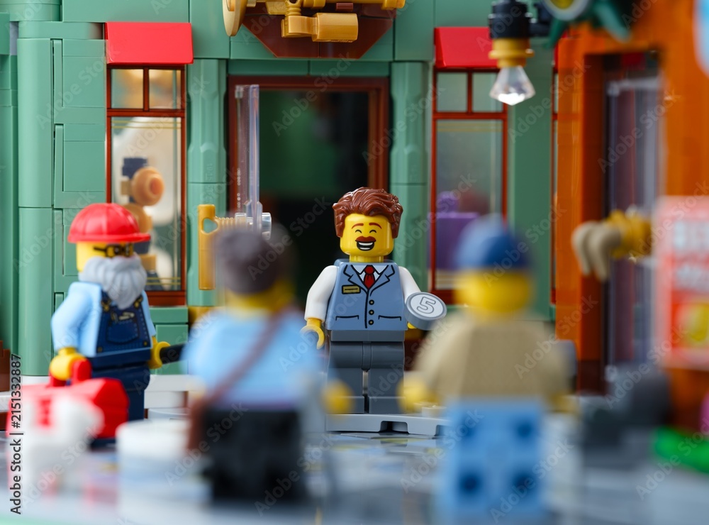 Fototapeta premium Tambov, Russian Federation - June 28, 2026 A smiling Lego man minifigure with a five value coin at the entrance of a store on a busy city street.