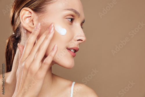 Young woman with brunette hair applies moisturizing cream on her cheek while looking sideways against a beige background with a calm and focused expression.