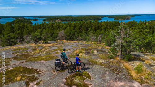 Father and son with loaded bicycles standing on rocky cliff overlooking archipelago islands during summer bikepacking trip on Saariston Rengastie Finland