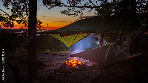 Hammock tents camping under pine trees with campfire and distant archipelago view during Nordic blue hour twilight