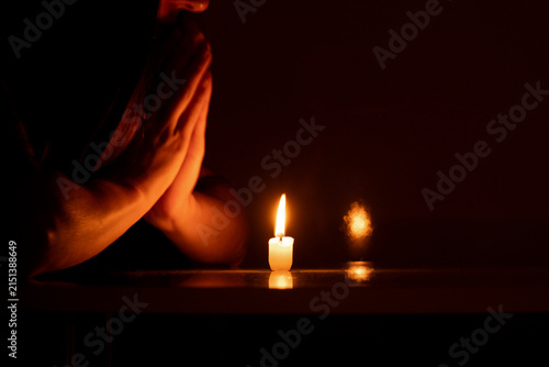 A woman with her hands folded in prayer before a burning candle in the dark as a symbol of faith, prayer, hope, fear