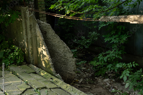 Foto Broken concrete fence wall with rebar exposed among wild green vegetation and debris in an abandoned overgrown area