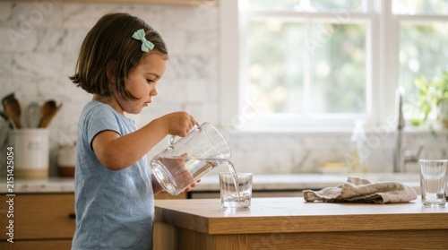 Child pours water from pitcher into glass in a bright kitchen, side-profile documentary moment with natural light and soft bokeh