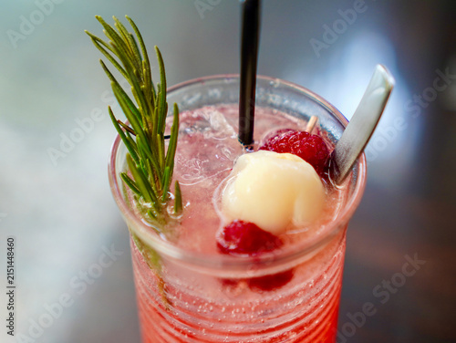 A glass of iced pink fruit mocktail garnished with lychee fruit, fresh berries, and rosemary on a reflective tabletop