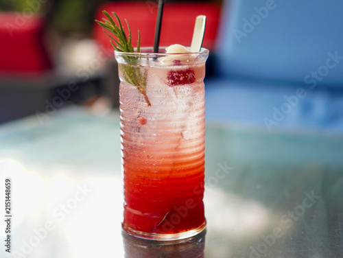 Vibrant red layered iced mocktail featuring fresh lychee fruit, berries, and rosemary garnish on a cafe table
