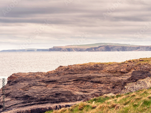 Rugged coastal cliffs with layered rock formations. Water pools between weathered stone surfaces. Green grass grows on cliff edge. Overcast sky above calm sea. Irish nature scenery, Rosses point.