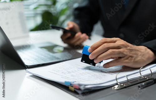 Businessman stamps a printed document at an office desk beside a laptop, symbolizing approval, verification, authorization, paperwork processing, compliance, and official administration.