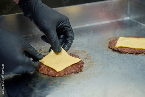 Obraz na płótnie Gloved Cook Hands Adding Melted Cheese Slice over Smash Burger Patty Cooking on