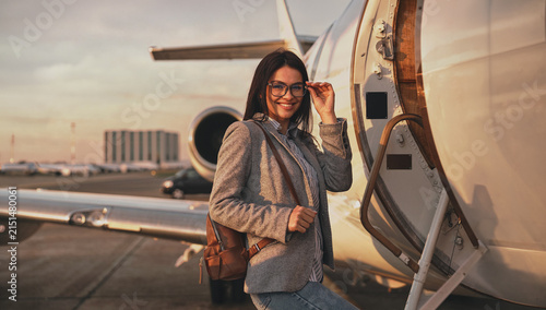 Fotografía A smiling woman, adjusting her glasses with one hand and gripping a tan leather backpack strap with the other, stands at the open stairway door of a private jet on a glowing golden hour tarmac
