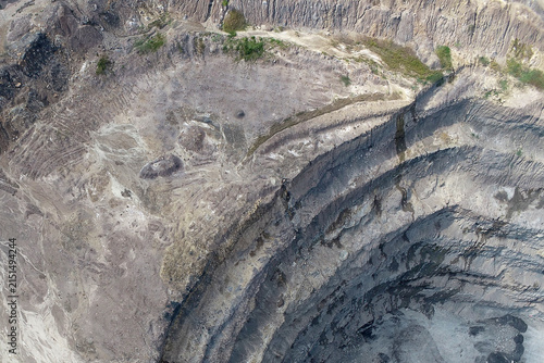 Fotografie Aerial oblique view of a deep open-pit mining highwall with curved terraced benches, exposed rock strata layers, and rugged excavated terrain under daylight