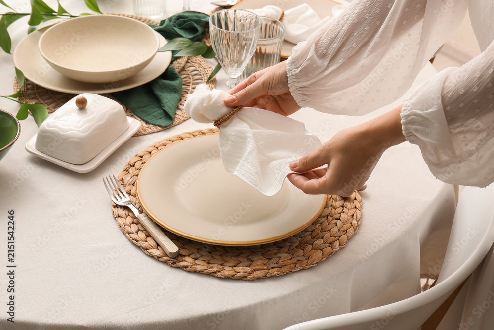 Fototapeta premium Womans hands setting a dining table with a white napkin and woven placemat for a meal or special occasion