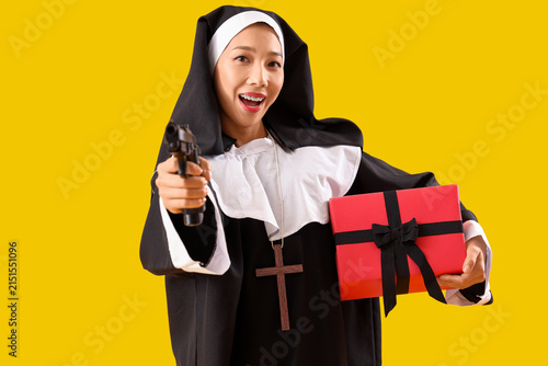 Young Asian woman dressed as nun with gun and Halloween gift on yellow backgr...