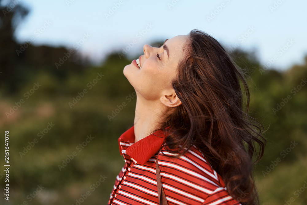 Obraz premium Young brunette woman looking up at the sky with a gentle smile outdoors. She wears a casual striped shirt capturing a peaceful lifestyle moment in the golden sunlight.