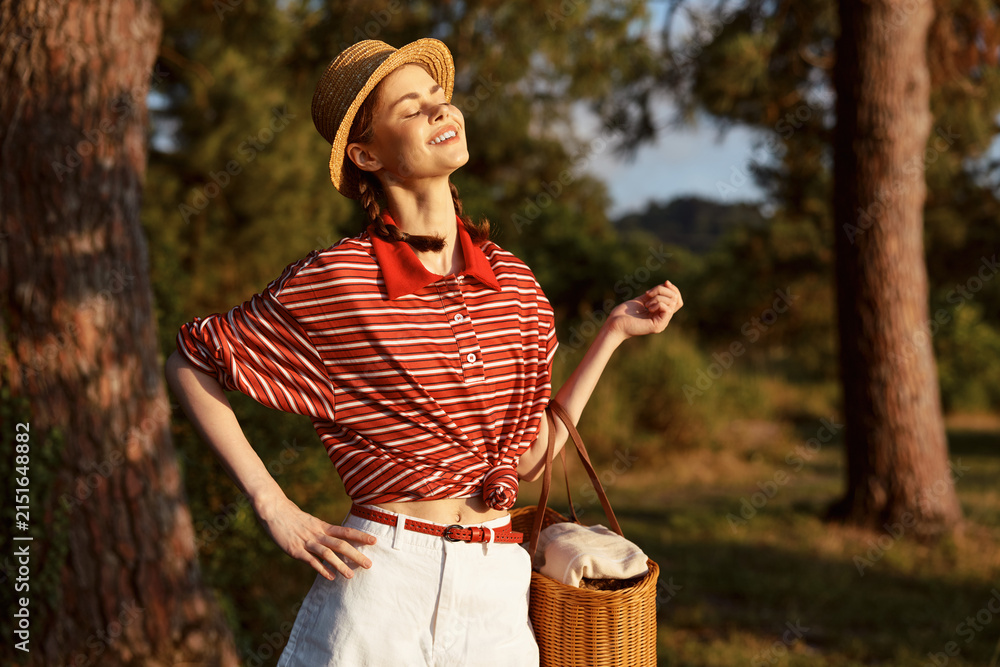 Obraz premium Young woman enjoying summer lifestyle outdoors in a garden. She wears a striped shirt, straw hat, and carries a wicker basket in the warm sun. Concept of leisure, vacation, and nature.
