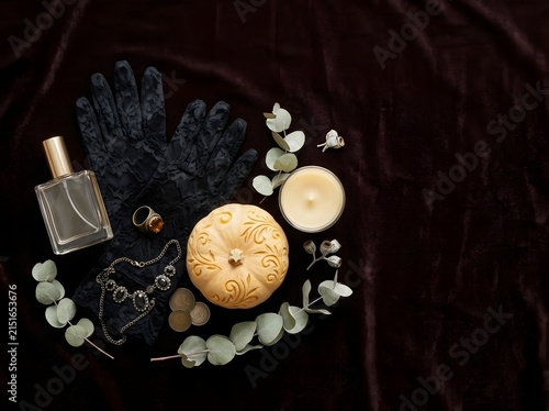 Flat lay composition of a luxury perfume bottle, black lace gloves, an intricately carved pumpkin, vintage jewelry, and a lit candle arranged on a dark brown velvet fabric background.
