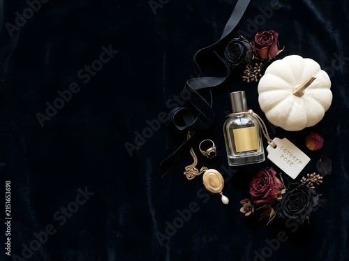 Flat lay composition on black velvet fabric with a glass perfume bottle, white mini pumpkin, vintage gold locket necklace, gold ring, dried dark red and black roses, and a ribbon.