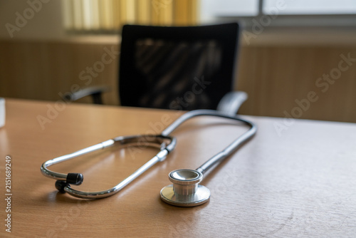 Hospital stethoscope on wooden table in empty doctor office with chair and soft light