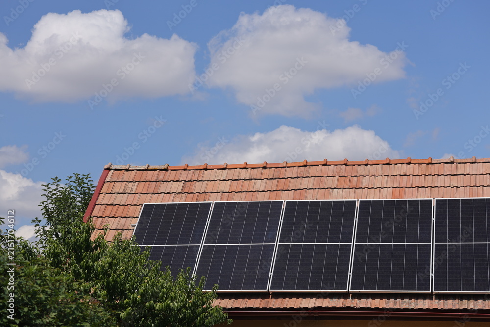 Fototapeta premium Solar panels on a red tiled roof beside a green fruit tree under a blue summer sky with white cumulus clouds