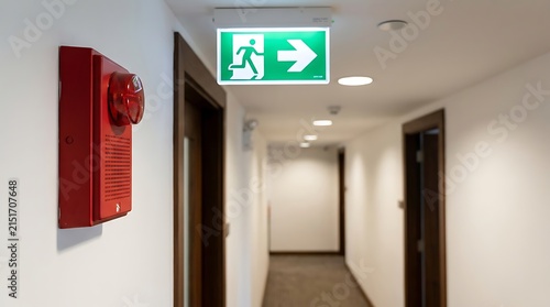 A red fire alarm strobe light mounted on a white wall in a long office corridor with a green illuminated emergency exit sign hanging above it
