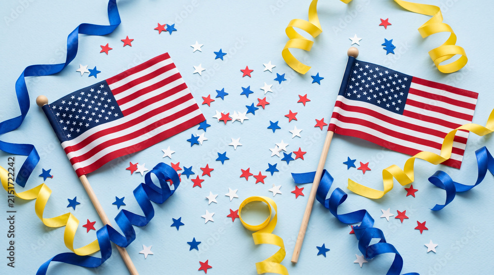 Obraz premium Small American flags with ribbons and star confetti on blue background.
