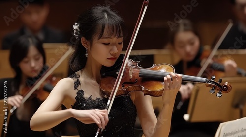 Asian female violinist performing on stage with orchestra in black dress