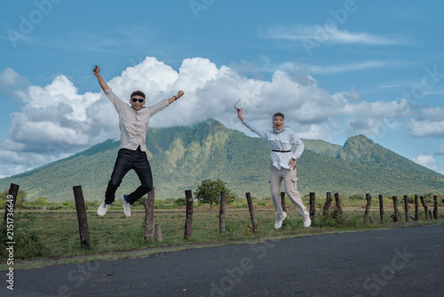 A joyful Asian couple laugh and jump on a rural road with a green Baluran mountain landscape and blue sky behind them, capturing adventure, freedom, travel and outdoor happiness during a scenic day tr