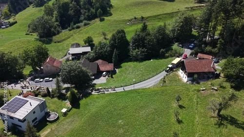 Aerial side view of a herd of cows descending a mountain road from the Swiss Alps, followed by farm tractors and cars waiting behind the animals.