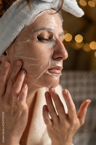 Middle aged woman applying anti wrinkle sheet mask to her face while performing an evening skincare routine at home.