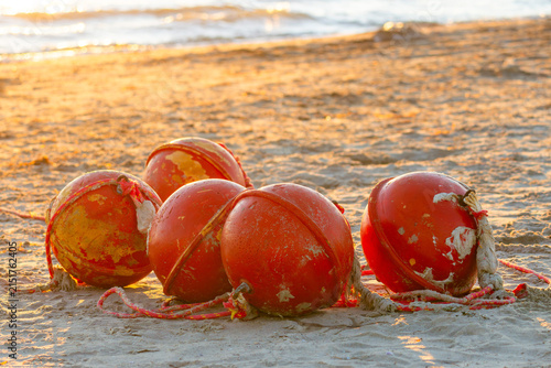 A group of worn orange fishing buoys with attached ropes lies on a sandy beach near the shoreline, illuminated by soft warm sunset light and gentle sea waves
