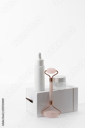 Minimalist facial care set featuring a rose quartz roller, white dropper bottle, and glass cream jar atop a sleek gift box. A clean, high-key studio shot perfect for luxury skincare branding.