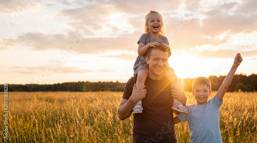 Father carries daughter on shoulders while son stands beside them in a golden field during sunset with trees in the background
