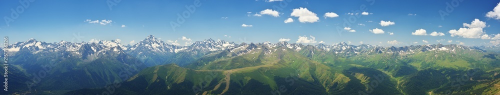 Fototapeta premium Extra wide panoramic landscape of a majestic mountain ridge with snowy peaks, glacier ice and green forested valleys under a blue sky with day haze.
