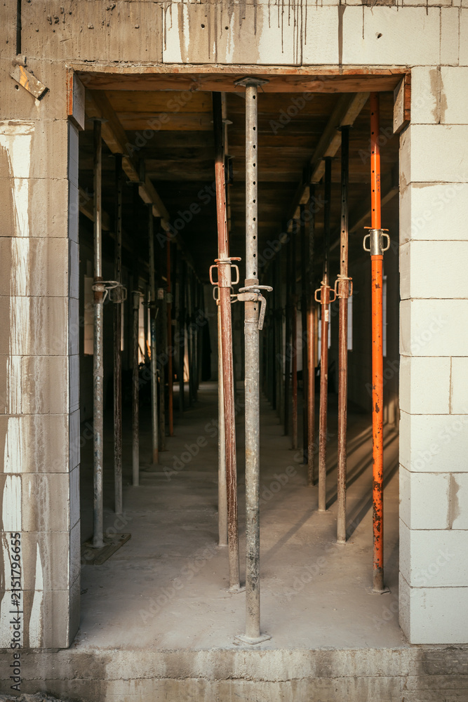 Fototapeta premium View through a block doorway of adjustable metal shoring jacks holding up a wooden ceiling structure on a construction site.