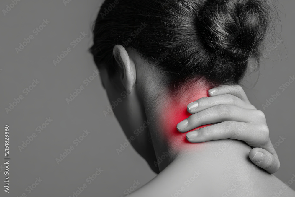 Fototapeta premium A person stands indoors, holding their neck with one hand, showing signs of discomfort. The focus is on the neck area which appears highlighted