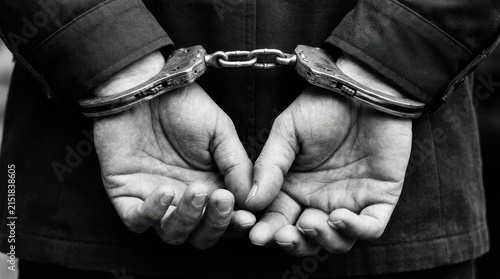 Detailed black and white close up of a suspect in metal handcuffs with hands behind their back during a police arrest, representing criminal justice and law enforcement for legal or editorial purpose.