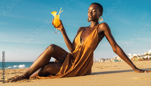 Woman in a golden dress relaxing on a sandy beach with a drink.