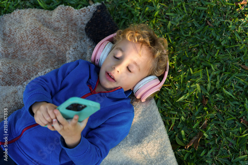 Toddler relaxing outdoors, listening to audio with pink headphones and smartphone on a blanket