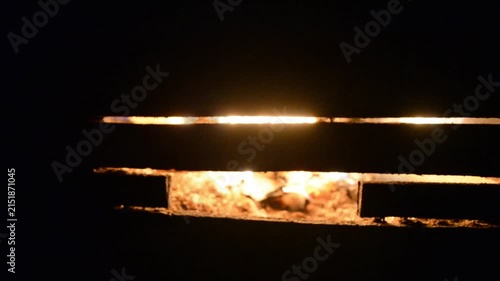 Fire glows behind dark wooden slats. Abstract view of burning logs inside a furnace or stove. Bright embers shine through horizontal gaps in the structure pattern.