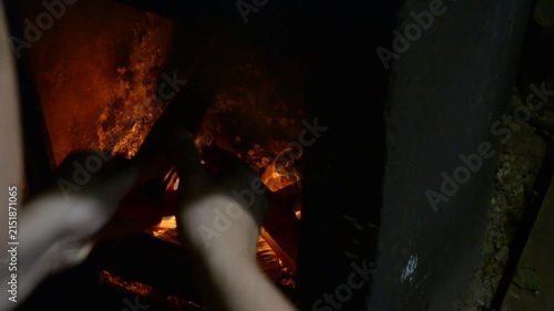Woman using tongs to add wood to a fireplace. Burning logs in a stone stove for home heating. Indoor scene of fire maintenance during a cold winter evening.