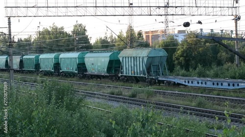 Freight train with green cargo hopper cars moving along railway tracks in countryside. Industrial logistics transport system on railroad during sunny summer day.
