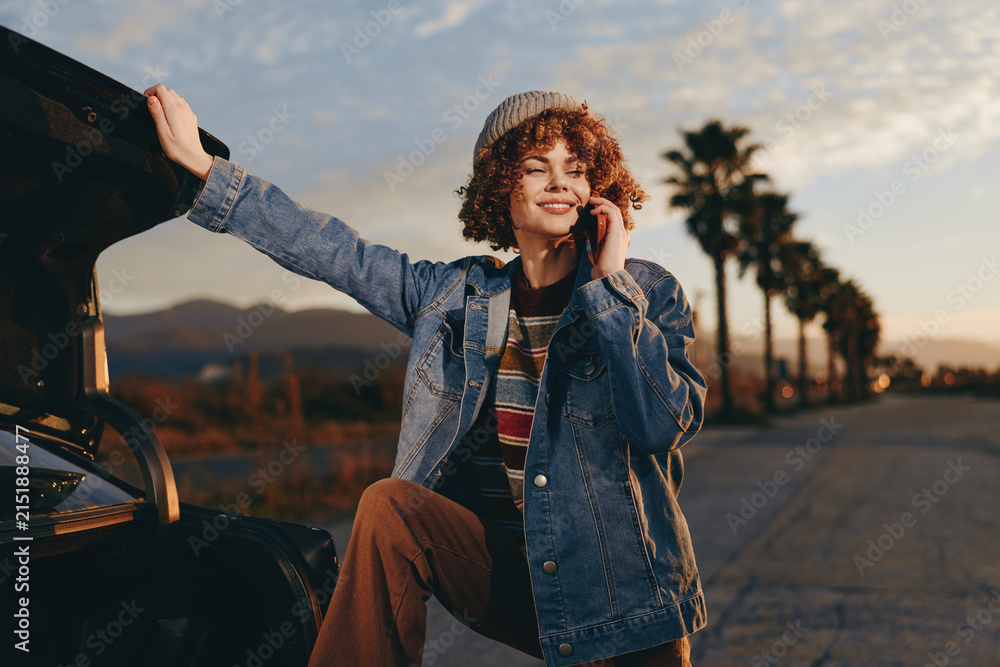 Obraz premium Smiling woman lifestyle outdoors near car at sunset, wearing denim jacket and curly hair, standing on street with warm evening light, relaxed travel pose and sunlit landscape.