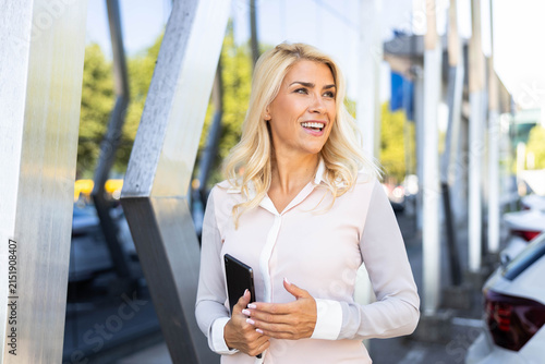 Confident blonde businesswoman laughing cheerfully, holding a digital tablet while walking in a modern city environment